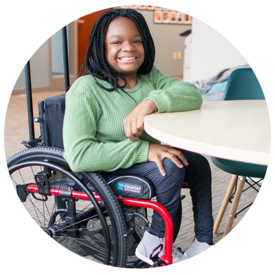 Young black girl with braids wearing a sweater and jean using a manual wheelchair smiling at the camera.